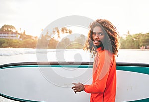 Portrait of black long-haired teen boy with surfboard ready for surfing with sunset backlight. He walking in Indian ocean waves.