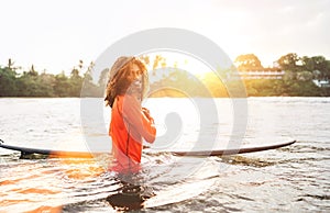 Portrait of black long-haired teen boy with surfboard ready for surfing with sunset backlight. He walking in Indian ocean waves.