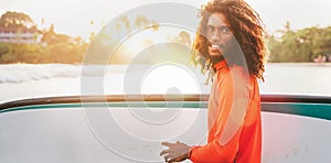 Portrait of black long-haired teen boy with surfboard ready for surfing with sunset backlight. He walking in Indian ocean waves.