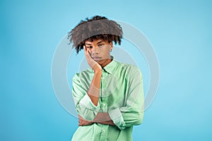 Portrait of black teen guy feeling bored, leaning on his hand, having dull day on blue studio background