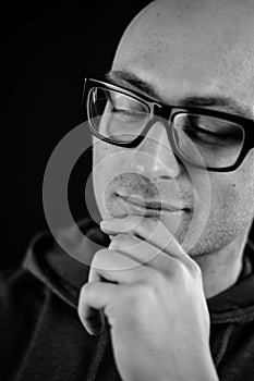 Portrait in black and white of a young bald man in black eyeglasses holding his chin with his hand while thinking looking down