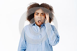 Portrait of black woman looking shocked and concerned at camera, gasps startled, stands over white studio background