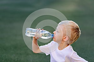 Portrait of a blond boy in a sports uniform sitting on a green lawn on a football field and drinking water from a bottle, sports