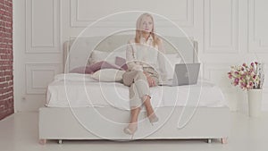 Portrait of a blond Caucasian businesswoman with grey eyes sitting with laptop on the bed and thinking. Young beautiful