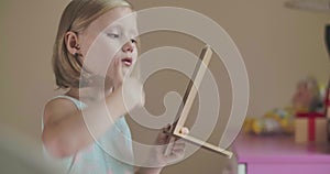 Portrait of blond Caucasian little girl looking at hand mirror and fixing her hair. Cute child in blue dress with pink