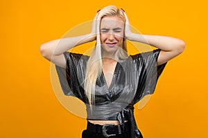 Portrait of a blond lady showing discontent covering her ears with hands on a yellow studio background