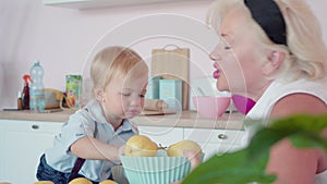 Portrait of blond little boy turning over basket with pears, cheerful Caucasian senior woman laughing. Charming child