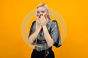 Portrait of a blond startled young woman covering her face with hands on a yellow studio background