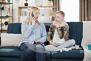Portrait of blond woman mother with thermometer and smartphone calling to doctor at home.