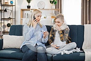 Portrait of blond woman mother with thermometer and smartphone calling to doctor at home.