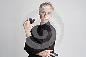 Portrait of blond woman with short hair wearing black shirt drinks takeaway coffee, using smartphone on white background.