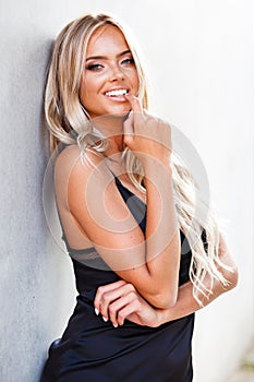 Portrait of blond young woman with beautiful makeup and hair in a classic black dress standing near grey wall. Fashion