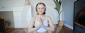 Portrait of blond young woman meditating, practise yoga, using yoga podcast to workout at home, sits on rubber mat