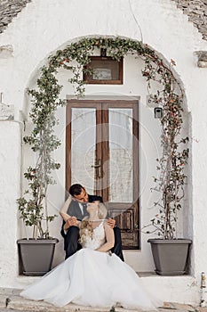 Portrait of blonde bride in white dress and brunet groom in suit kissing and sitting near old city house with arch from