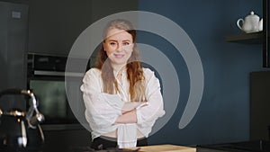 Portrait of blonde caucasian woman crossing arms and smiling in her kitchen. Young cheerful adult female folding hands