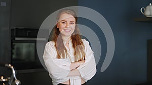 Portrait of blonde caucasian woman crossing arms and smiling in her kitchen. Young cheerful adult female folding hands