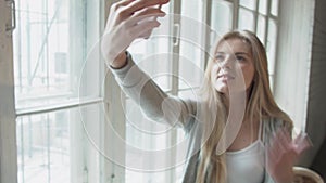 Portrait of a blonde doing selfie by the window. Young beautiful woman sends an air kiss through the phone.