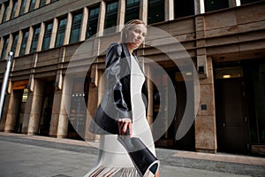 Portrait of blonde female fashion model in leather blazer jacket and white dress looking into the camera, walking in