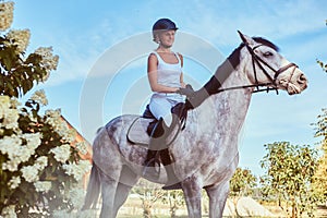 Portrait of blonde female riding a dapple gray horse near lilac bushes in garden.