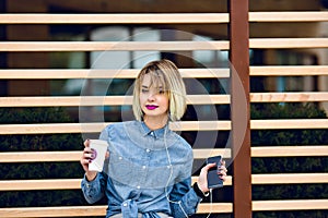 A portrait of a blonde girl with a forelock and bright pink lips wearing blue jeans shirt and looking straight, holding
