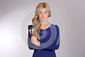 Portrait of a blonde makeup artist girl with blonde hair in a blue dress with face brushes in the studio room, with makeup and