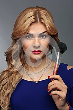 Portrait of a blonde makeup artist girl with blonde hair in a blue dress with face brushes in the studio room, with makeup and