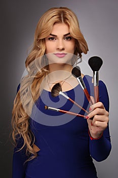 Portrait of a blonde makeup artist girl with blonde hair in a blue dress with face brushes in the studio room, with makeup and