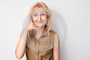 Portrait of blonde mature woman pretends calling someone, makes call me sign with fingers