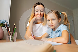 Portrait of blonde primary daughter doing homework with young mother sitting at home table by window, looking at camera.