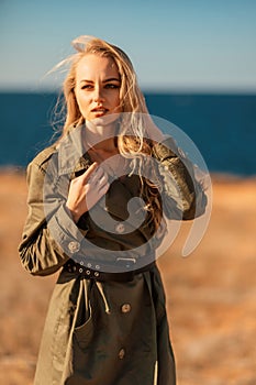 Portrait blonde sea cape. Calm young blonde in a khaki raincoat stands on the seashore