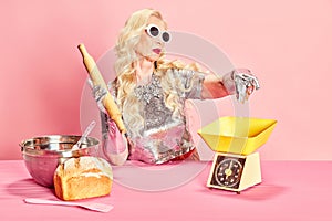 Portrait of blonde, senior, beautiful lady, woman in stylish silver dress and sungasses cooking bread against pink