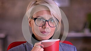 Portrait of blonde short-haired businesswoman in glasses invitingly watching into camera and drinking coffee in office.