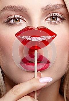 Portrait of blonde woman closes by candy her nose. Red female lips shape lollipop. Sweet tooth concept