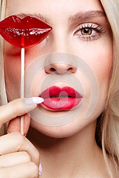 Portrait of blonde woman closing by candy her eye.. Red female lips shape lollipop. Sweet tooth concept