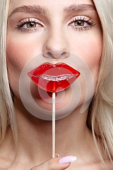 Portrait of blonde woman  kissing candy. Red female lips shape lollipop. Sweet tooth concept