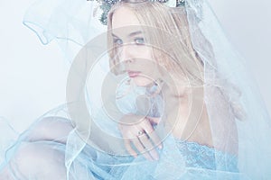 Portrait of a blonde woman with a wreath on her head and a blue delicate light transparent dress. Big blue eyes and beautiful skin