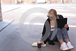 Portrait of a blonde working on a laptop while sitting on the pavement in the shade. Makes notes in the diary. Nearby is a glass