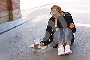 Portrait of a blonde working on a laptop while sitting on the pavement in the shade. Makes notes in the diary. Nearby is a glass