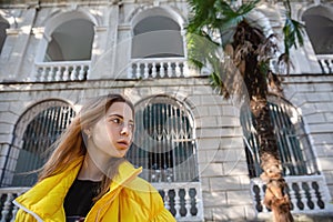 Portrait of blonde in yellow jacket against the background of an old building and palm tree. Travel and urban tourism.