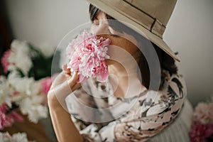 Portrait of boho girl smelling peony at pink and white peonies on rustic wooden floor. Stylish hipster woman in bohemian dress