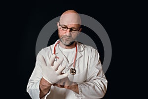 Portrait of a bold slim doctor on a dark background putting on a white rubber glove. Caucasian male in his 40s, grey beard,