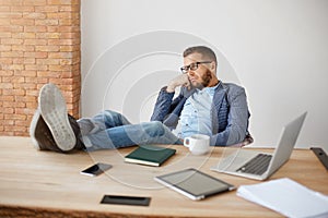 Portrait of bored adult caucasian unshaven male company manager in glasses and blue suit sitting with legs on table with