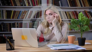 Portrait of bored blonde middle-aged caucasian woman typing using laptop in office on bookshelves background.