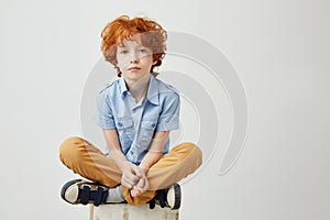 Portrait of bored little kid with red hair and freckles sitting on box with unhappy expression, being tired of waiting