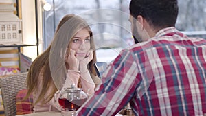 Portrait of bored pretty Caucasian girl sitting with boyfriend or husband in restaurant as her spouse using smartphone