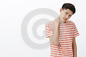 Portrait of bothered uneasy young asian man in striped t-shirt unwilling to do something rubbing neck tilting head and photo