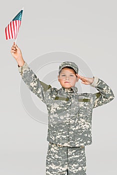 portrait of boy in camouflage clothing saluting while holding american flagpole in hand