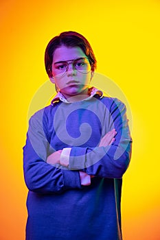 Portrait of boy, child in blue sweater and glasses posing with serious expression isolated over yellow studio background