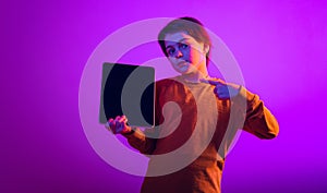 Portrait of boy, child pointing at tablet isolated over purple background in neon light