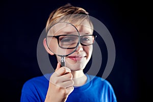 A portrait of a boy with eyeglasses looks through a magnifying glass in front of dark background. Children and education concept